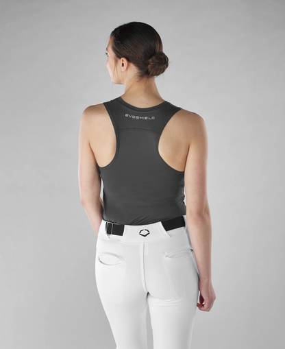 A woman with her hair in a bun, wearing an EvoShield Women's Racerback Chest Guard Sleeveless Shirt and white pants with a black belt, stands facing away against a plain grey background.
