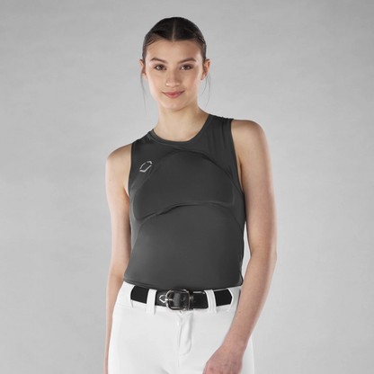 A woman with dark hair in a ponytail models the EvoShield Women's Racerback Chest Guard Sleeveless Shirt (WB5747701 / WB5747601) by EvoShield, paired with white pants and a black belt against a plain gray background, smiling slightly.
