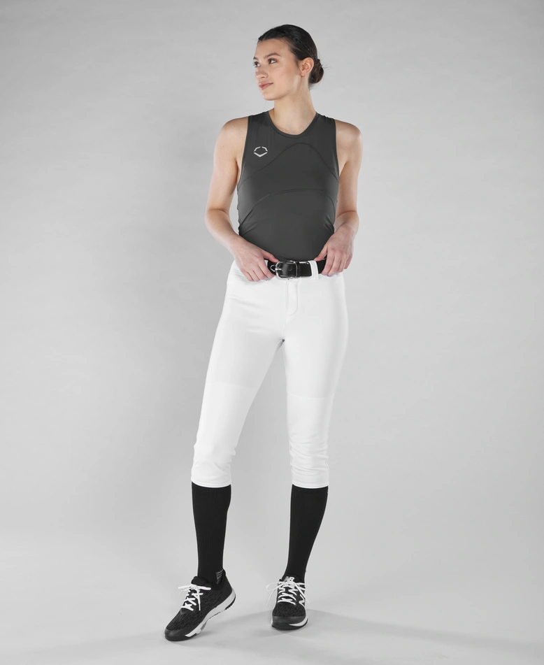 Against a gray background, a person wears the EvoShield Women's Racerback Chest Guard Sleeveless Shirt (WB5747701/WB5747601), white baseball pants, black belt, black knee-high socks, and black cleats, looking to the side with hands on hips.