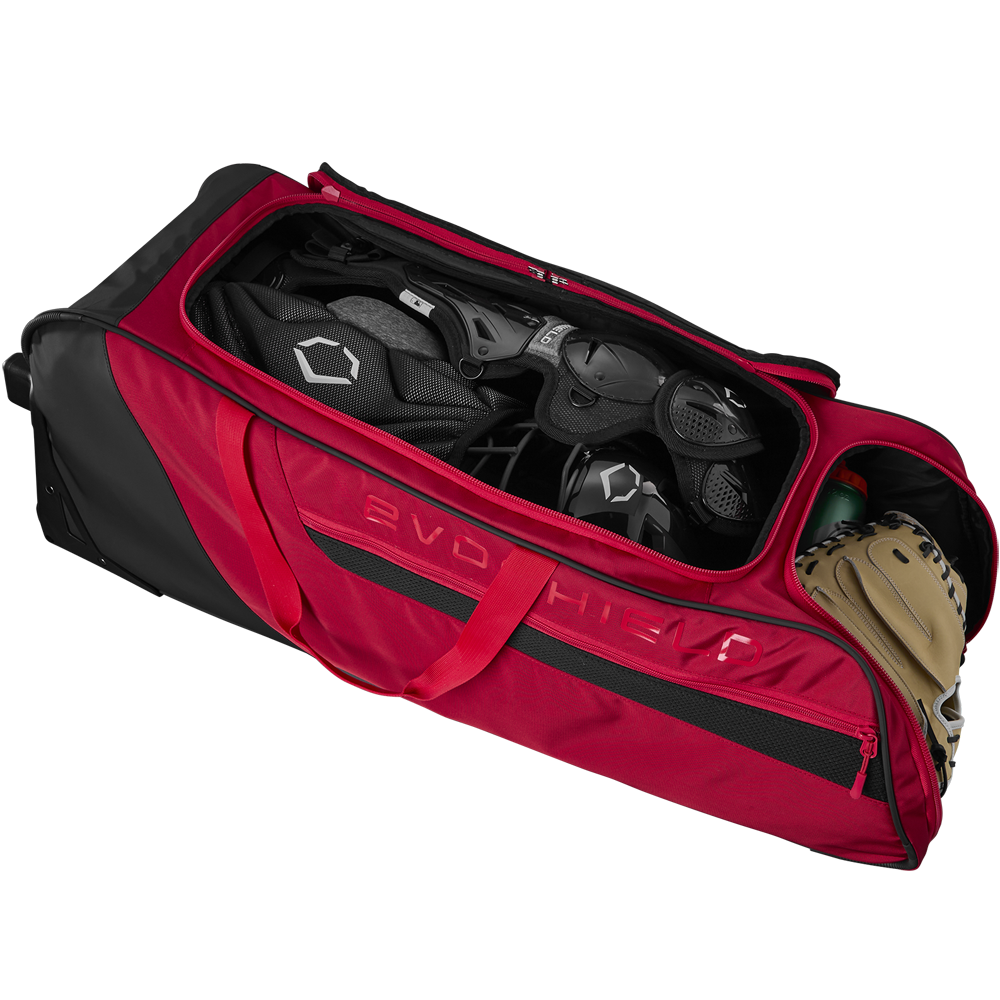 The EvoShield Takeover Wheeled Bag (WB57572) by EvoShield is unzipped, showing spacious bat sleeves and baseball equipment inside, including a helmet, catcher’s gear, and a tan glove.