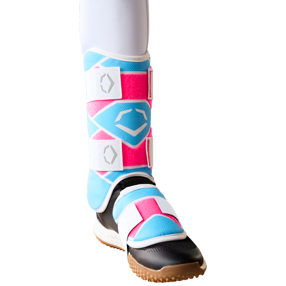 A lower leg wears the EvoShield SRZ-2 Youth Batter's Leg Guard (WB57646) by EvoShield, featuring pink, blue, and white geometric patterns, secured with white Velcro straps and a black and brown sole—resembling youth baseball gear.