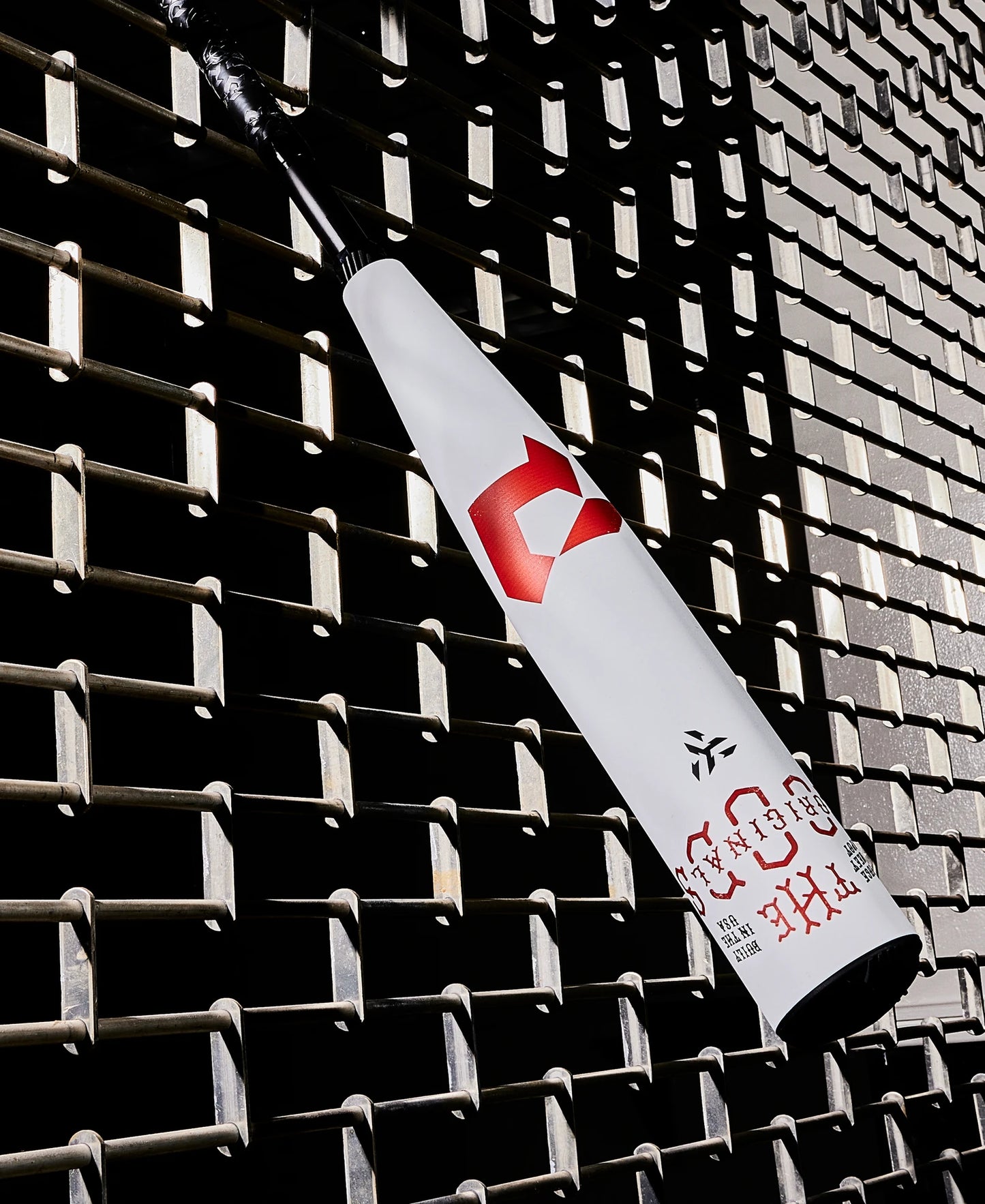 A white 2025 DeMarini The Goods (-8) 2 3/4" USSSA Baseball Bat (WBD2536010) with red logos and a black handle leans diagonally against a textured metal grid wall, its X14 Alloy Barrel casting distinct shadows.