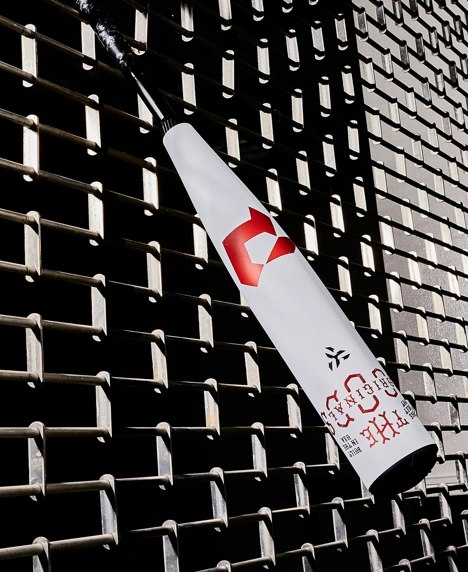 A white 2025 DeMarini The Goods (-8) 2 3/4" USSSA Baseball Bat (WBD2536010) with red logos and a black handle leans diagonally against a textured metal grid wall, its X14 Alloy Barrel casting distinct shadows.