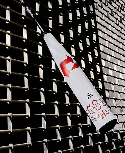 A white 2025 DeMarini The Goods (-8) 2 3/4" USSSA Baseball Bat (WBD2536010) with red logos and a black handle leans diagonally against a textured metal grid wall, its X14 Alloy Barrel casting distinct shadows.