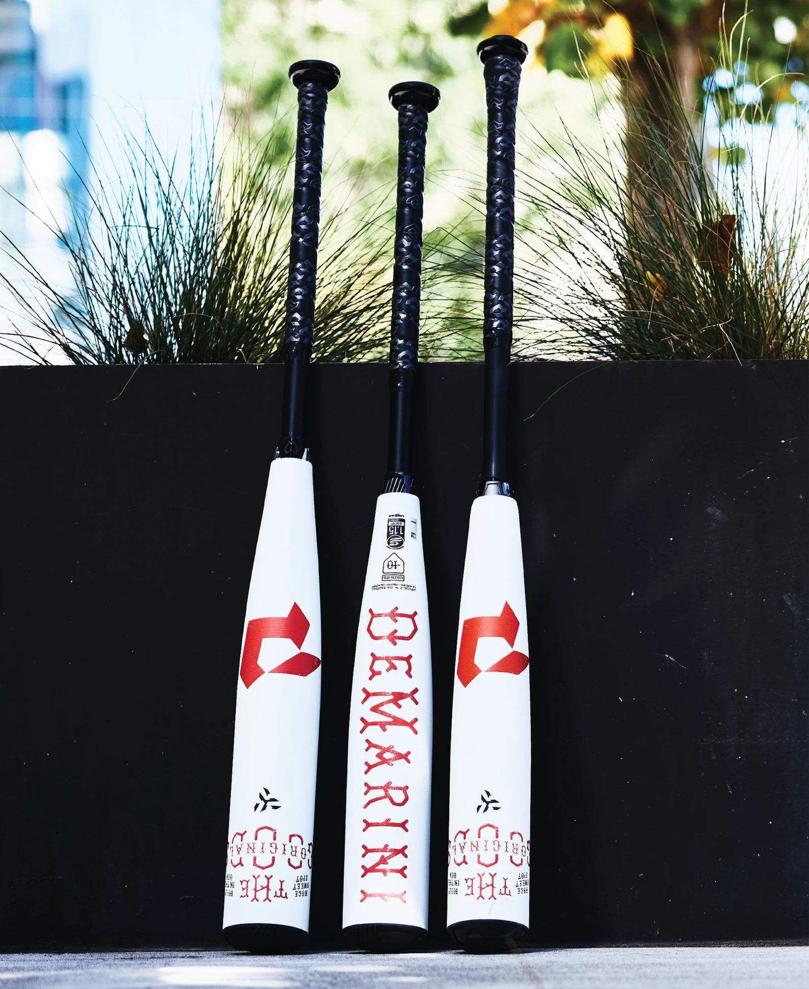 Three DeMarini The Goods 2025 (-8) 2 3/4" USSSA Baseball Bats (WBD2536010), white with black handles and red branding, stand upright by a planter with green plants in a blurred outdoor setting. Features X14 Alloy Barrel.