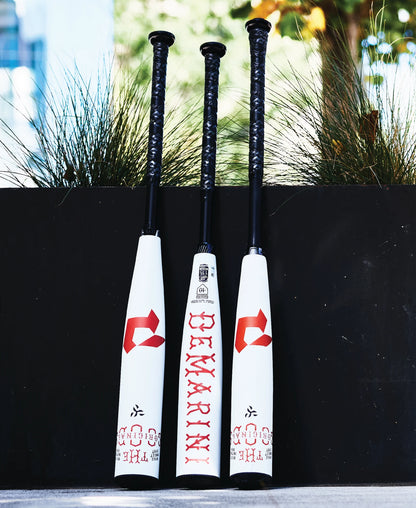 Three DeMarini The Goods 2025 (-8) 2 3/4" USSSA Baseball Bats (WBD2536010), white with black handles and red branding, stand upright by a planter with green plants in a blurred outdoor setting. Features X14 Alloy Barrel.