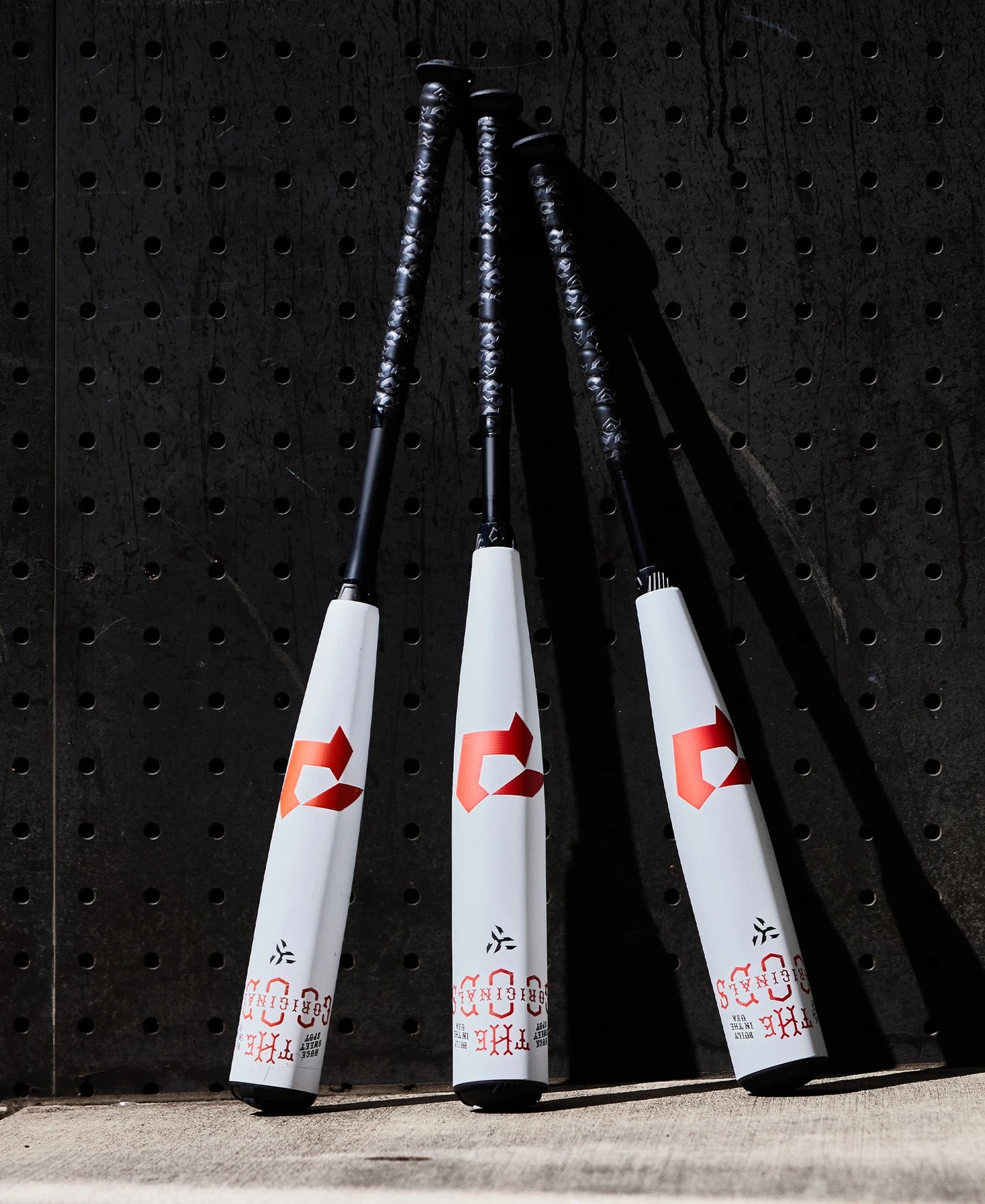 Three 2025 DeMarini The Goods (-8) USSSA baseball bats with white barrels, black handles, and red logos lean upright against a black pegboard wall, each featuring a powerful X14 Alloy Barrel.