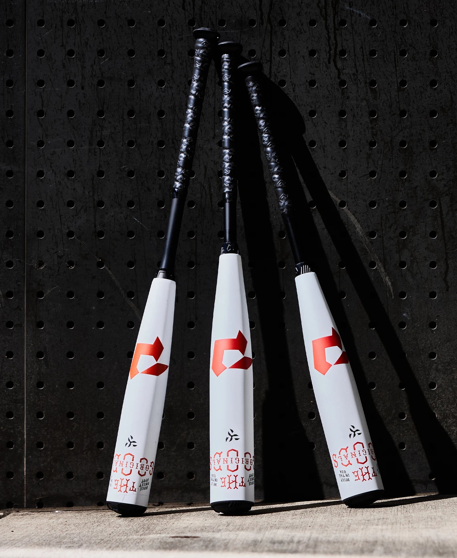 Three 2025 DeMarini The Goods (-8) USSSA baseball bats with white barrels, black handles, and red logos lean upright against a black pegboard wall, each featuring a powerful X14 Alloy Barrel.
