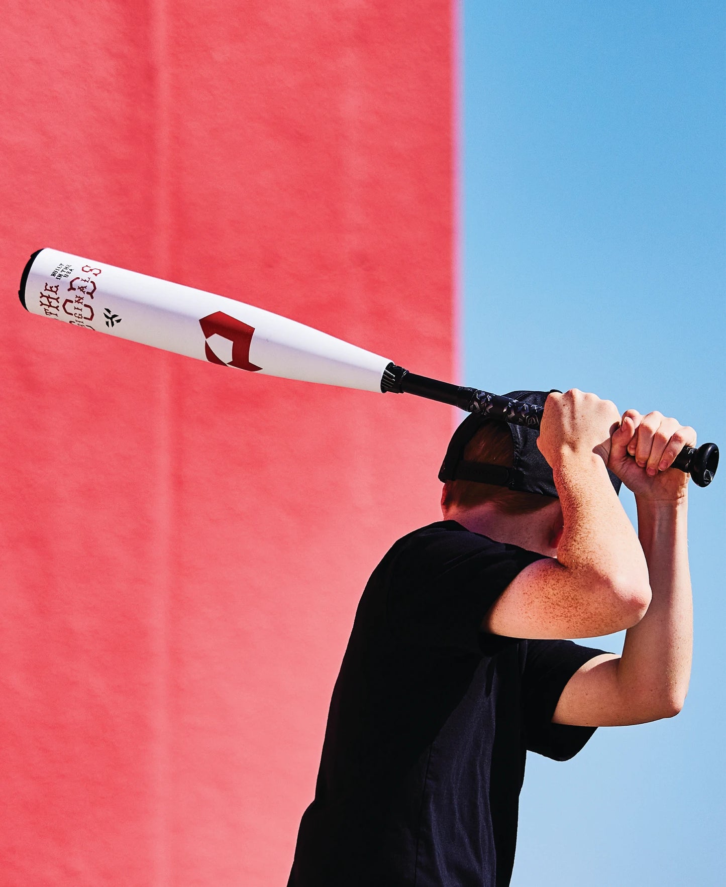 A person in a black shirt holds a DeMarini 2025 The Goods (-8) 2 3/4" USSSA Baseball Bat (WBD2536010) over their shoulder, standing before a red wall under a blue sky, with their face partly hidden by both the cap and the bat.