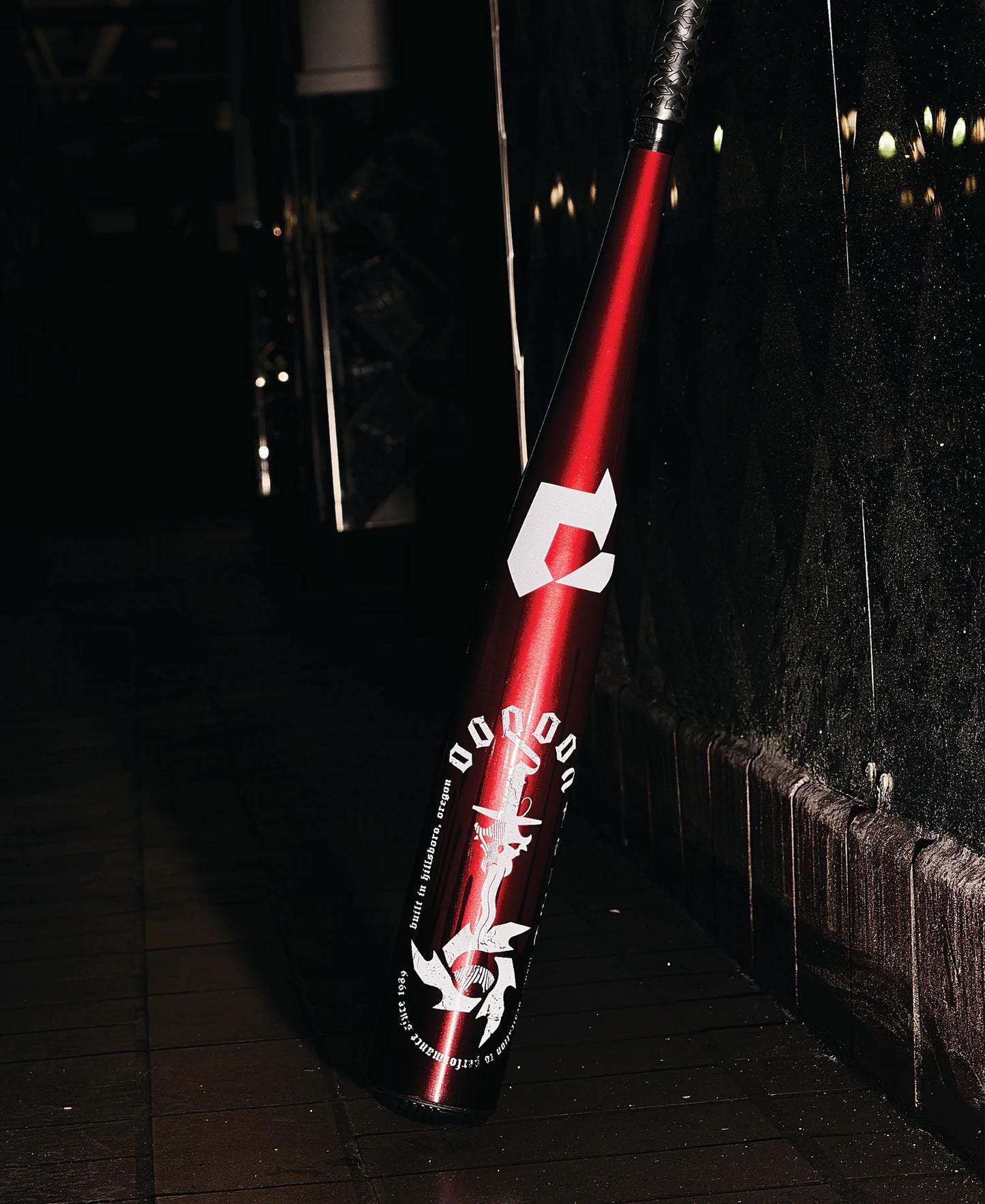 A 2025 DeMarini Voodoo One (-8) 2 3/4" USSSA Baseball Bat (WBD2539010) with a red metallic finish and white graphics leans against a tiled wall on a dark, wet sidewalk, featuring DeMarini’s X14 Alloy Barrel for peak performance.
