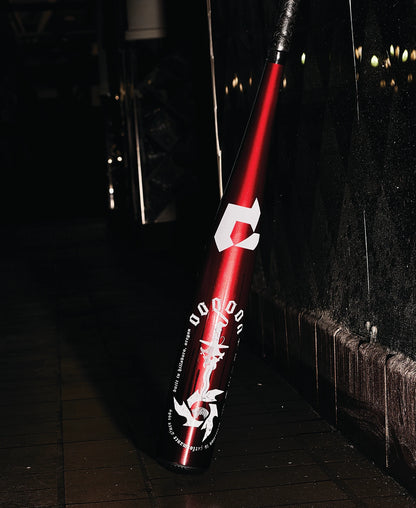 A 2025 DeMarini Voodoo One (-8) 2 3/4" USSSA Baseball Bat (WBD2539010) with a red metallic finish and white graphics leans against a tiled wall on a dark, wet sidewalk, featuring DeMarini’s X14 Alloy Barrel for peak performance.
