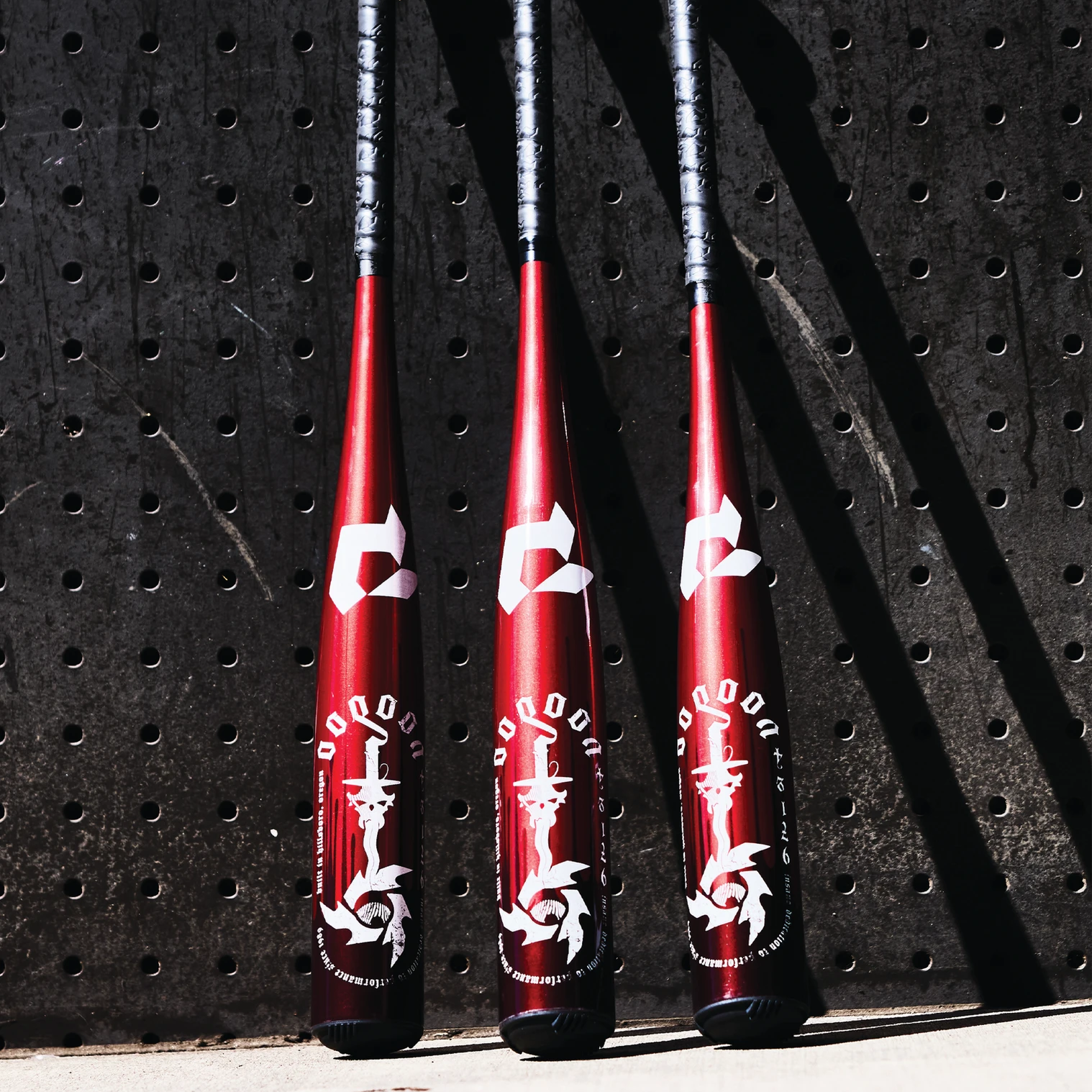 Three red baseball bats with white decorative designs stand upright against a dark, perforated metal wall, casting shadows on the ground.