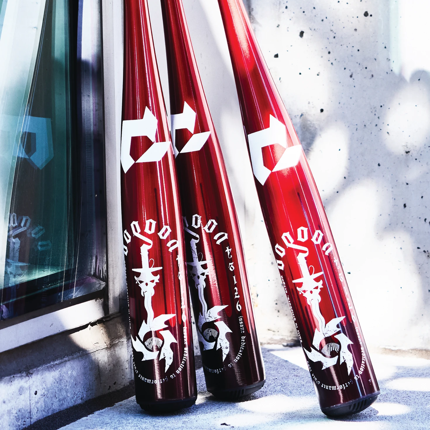 Three glossy red baseball bats with bold white Japanese characters and designs lean against a concrete wall beside a glass window, casting reflections and shadows on the ground.