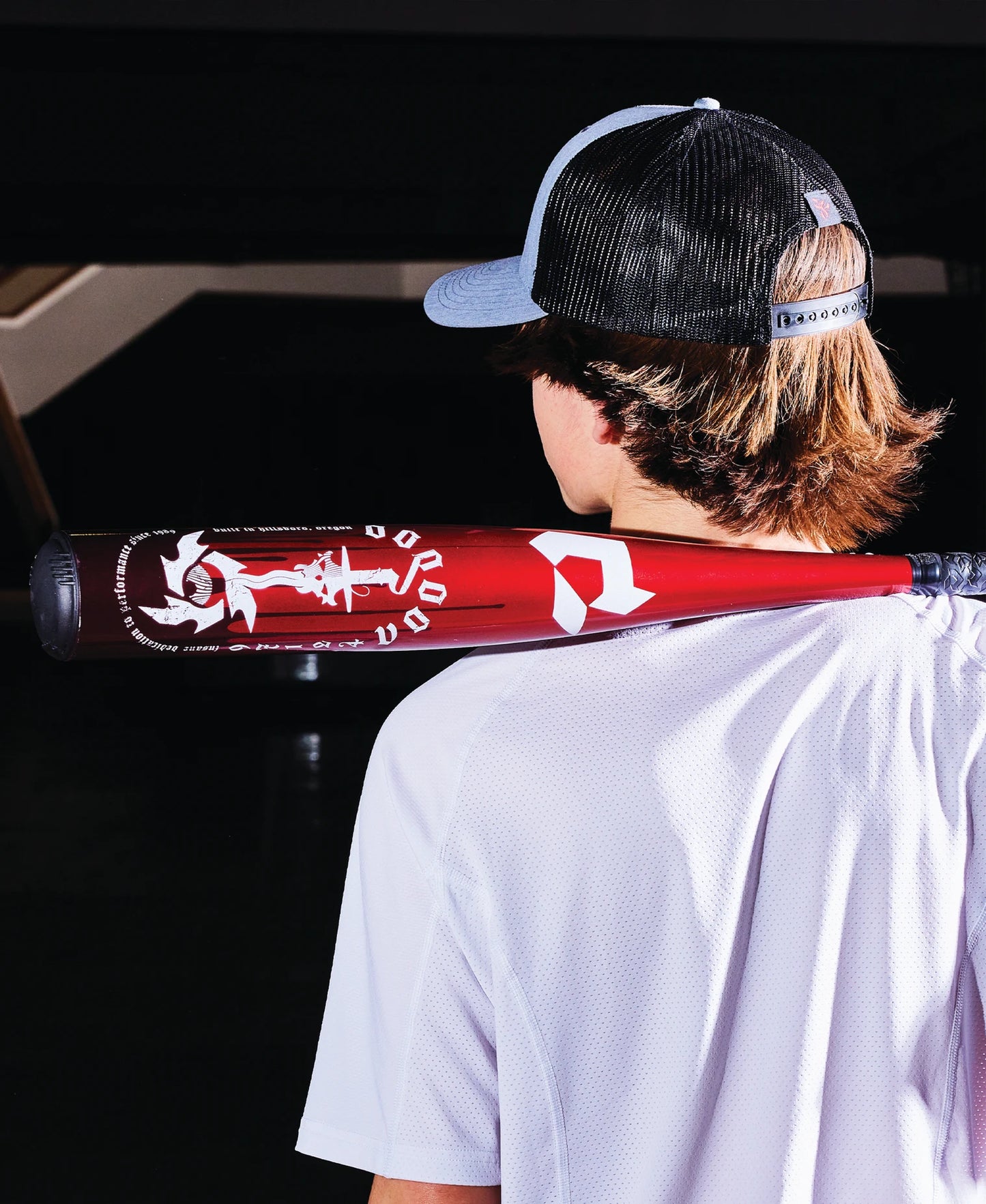 A person in a black and gray cap and white shirt stands with their back to the camera, resting a red 2025 DeMarini Voodoo One (-8) 2 3/4" USSSA Baseball Bat (WBD2539010 DEMO) with white graphics on their shoulder.
