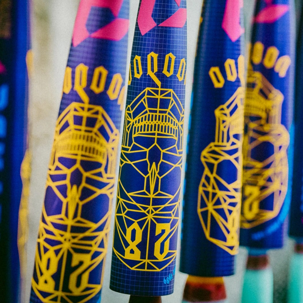 Close-up of colorful baseball bats featuring bold, geometric yellow designs and numbers on a blue background, with pink and blue accents, hanging in a row.