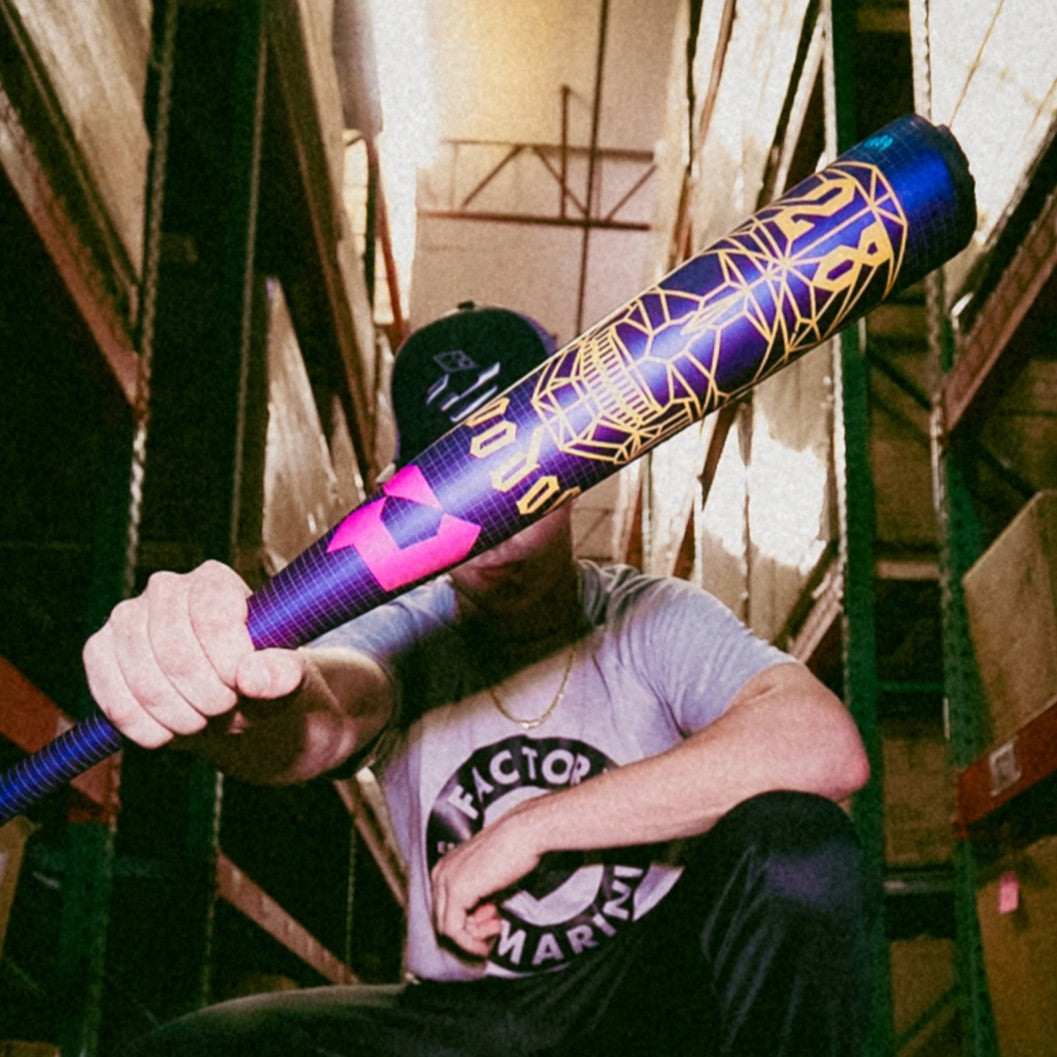 A person in a warehouse points a colorful, geometric-patterned baseball bat toward the camera, wearing a T-shirt, chain necklace, and a cap that partly obscures their face.