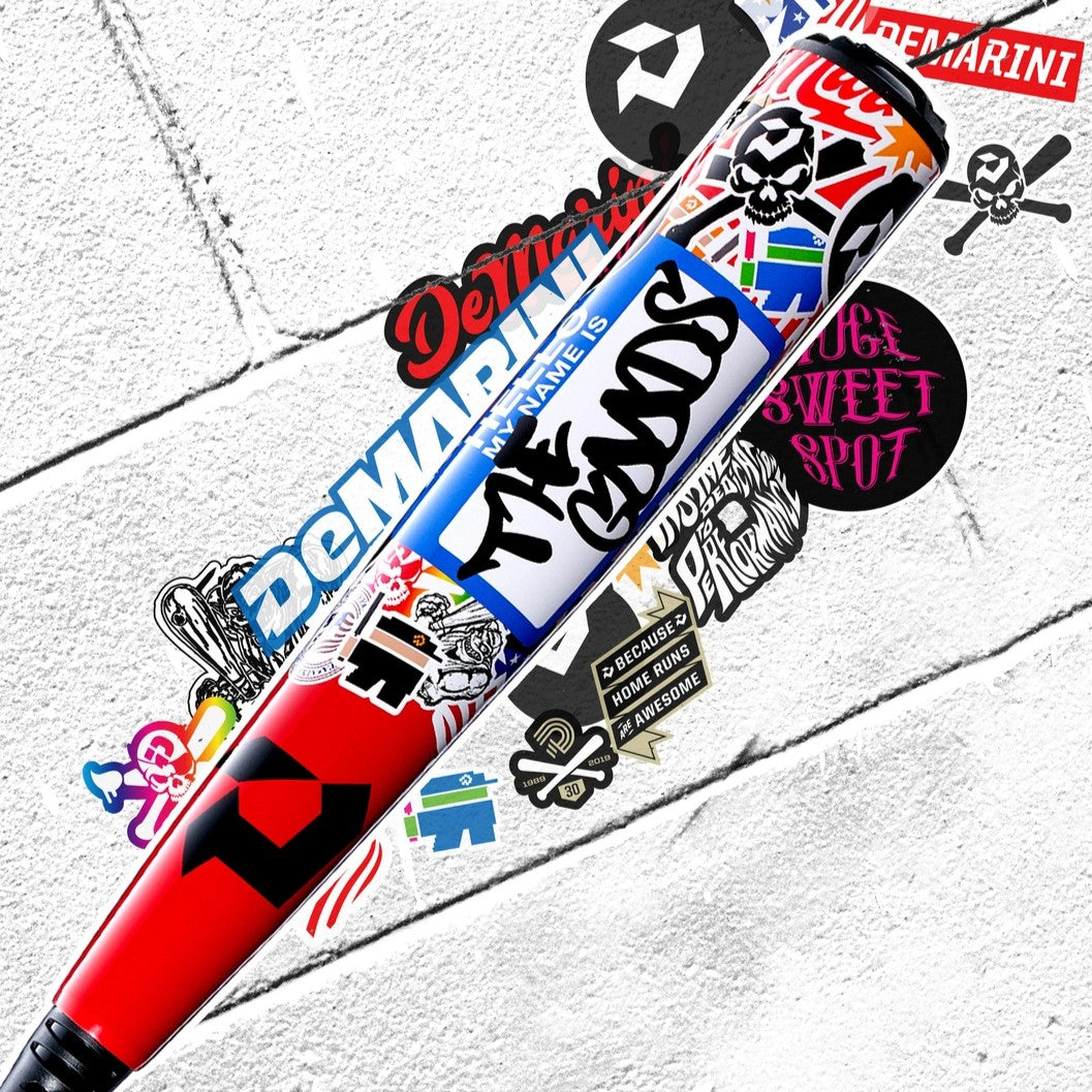 A baseball bat covered in colorful, overlapping stickers lies on a white concrete surface with more stickers, including logos and graffiti-style designs, scattered around it.