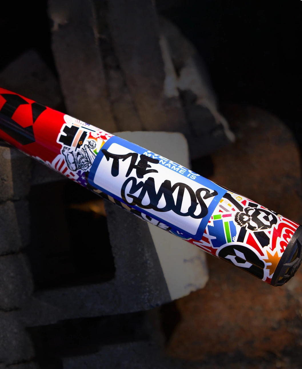 The 2026 DeMarini The Goods Sticker Bomb (-3) BBCOR Baseball Bat (WBD2653010) features vibrant stickers, including a bold blue "THE GOODS" label, set against a backdrop of concrete blocks and sand.