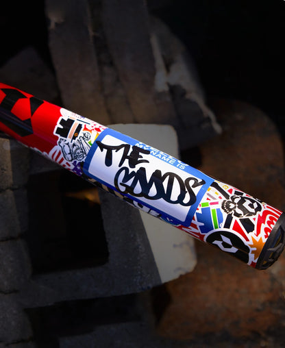 The 2026 DeMarini The Goods Sticker Bomb (-3) BBCOR Baseball Bat (WBD2653010) features vibrant stickers, including a bold blue "THE GOODS" label, set against a backdrop of concrete blocks and sand.