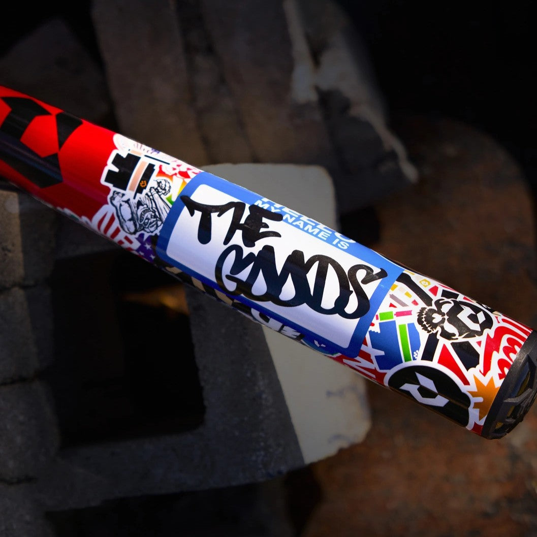 A colorful object covered in various stickers, including a prominent blue name tag sticker with THE GOODS written in bold black marker, resting on stacked concrete blocks.