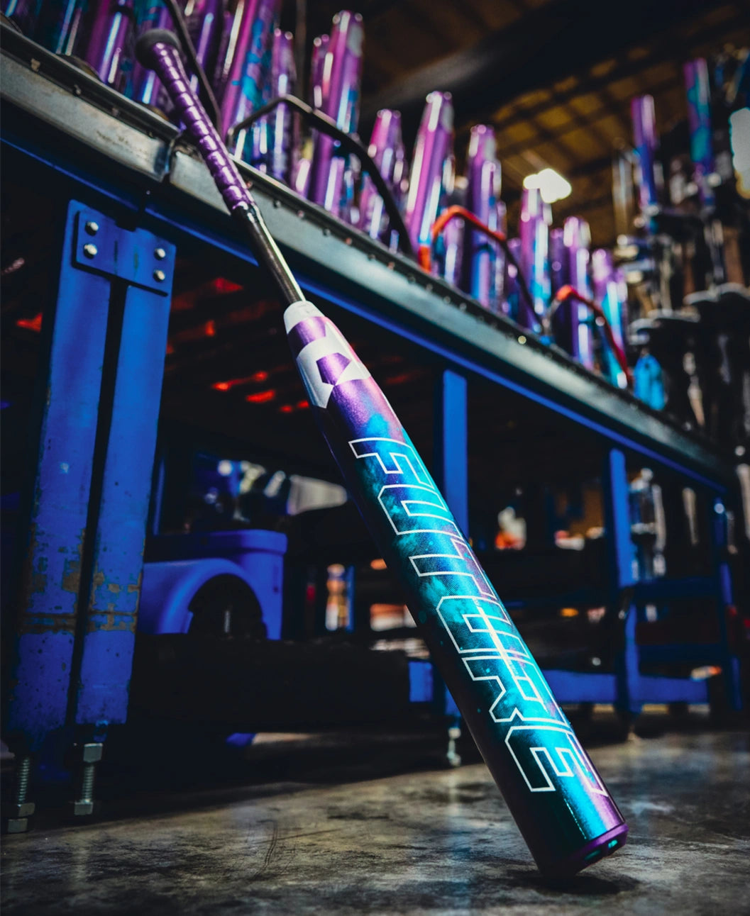 A 2026 DeMarini Future (-10) Fastpitch Softball Bat (WBD2674010), featuring a metallic purple, teal, and white design labeled FUTURE, leans against a blue rack in a factory, surrounded by other colorful DeMarini bats with large sweet spots.