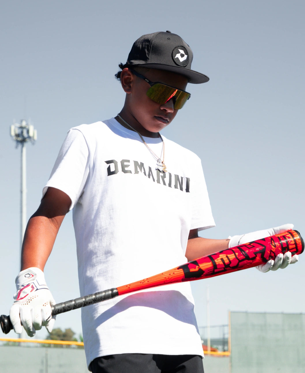 A young person wearing sunglasses, a black cap, white DeMarini t-shirt, and gloves stands outdoors under a clear sky, holding the 2027 DeMarini Exile (-11) 2 5/8″ USA Baseball Bat: WBD2719010 from DeMarini.