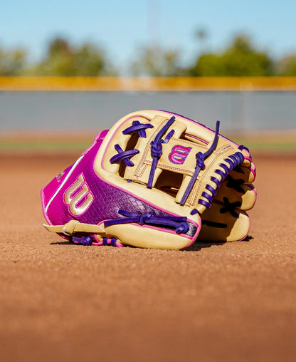 A Wilson A2000 1720SS 11.5″ Baseball Glove (WBG0009401RT115) in purple and tan rests on the dirt of a baseball field, with a blurred fence and blue sky in the background, highlighting Wilson’s classic infielder’s glove design.