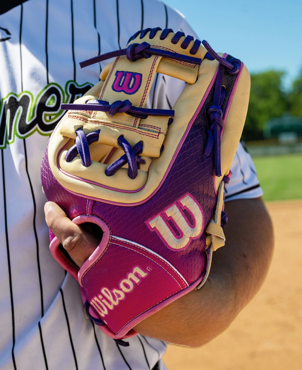 On a sunny baseball field, a person in a striped baseball jersey holds the Wilson A2000 1720SS 11.5″ Baseball Glove (WBG0009401RT115) by Wilson—featuring a pink and cream design with purple laces—designed for infielders.