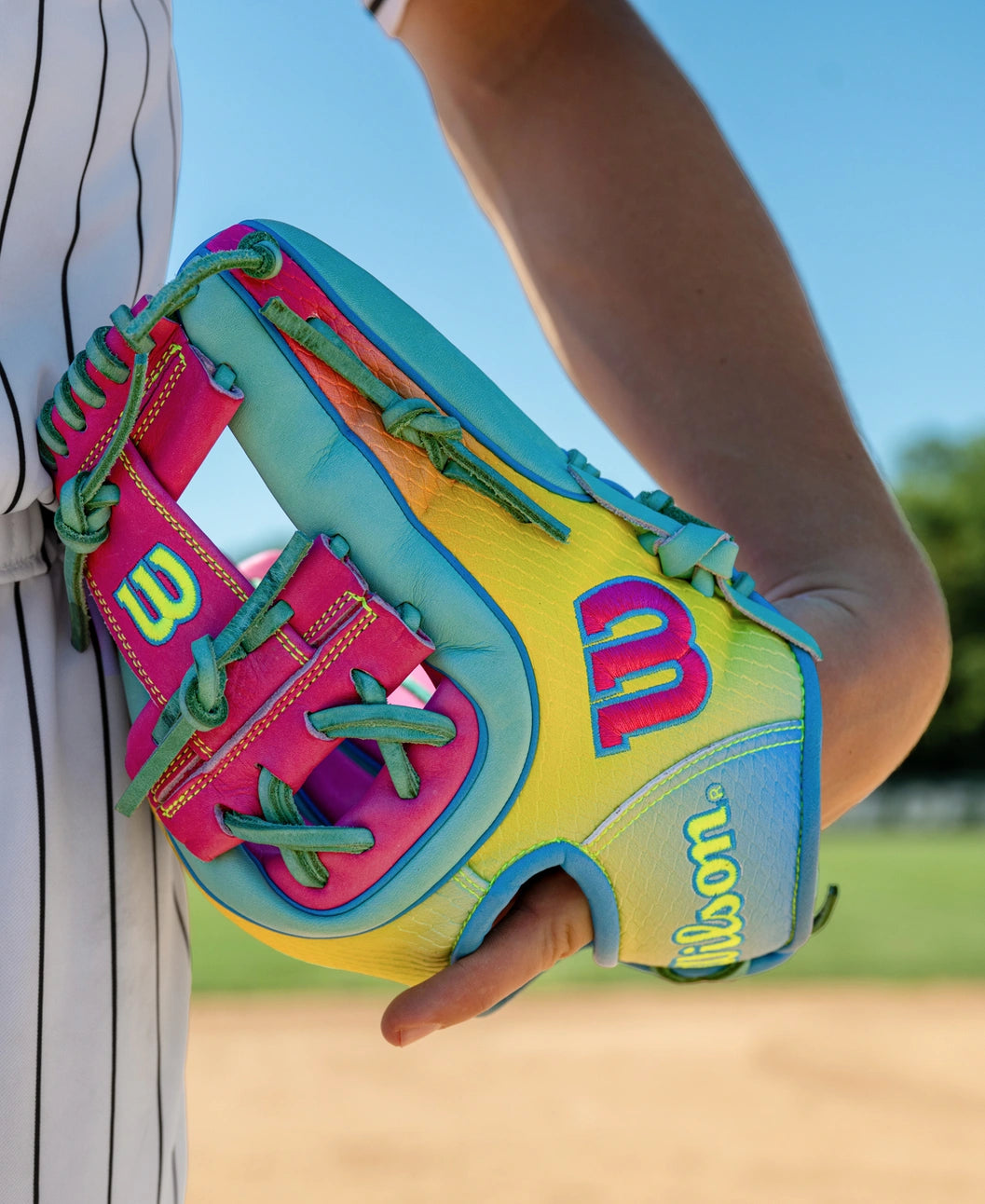 A person in a striped baseball uniform holds a colorful Wilson A2000 1727SS 11.5″ Baseball Glove: WBG0009501RT1175, featuring vibrant blue, pink, and yellow SuperSnakeSkin sections, on a sunny baseball field.