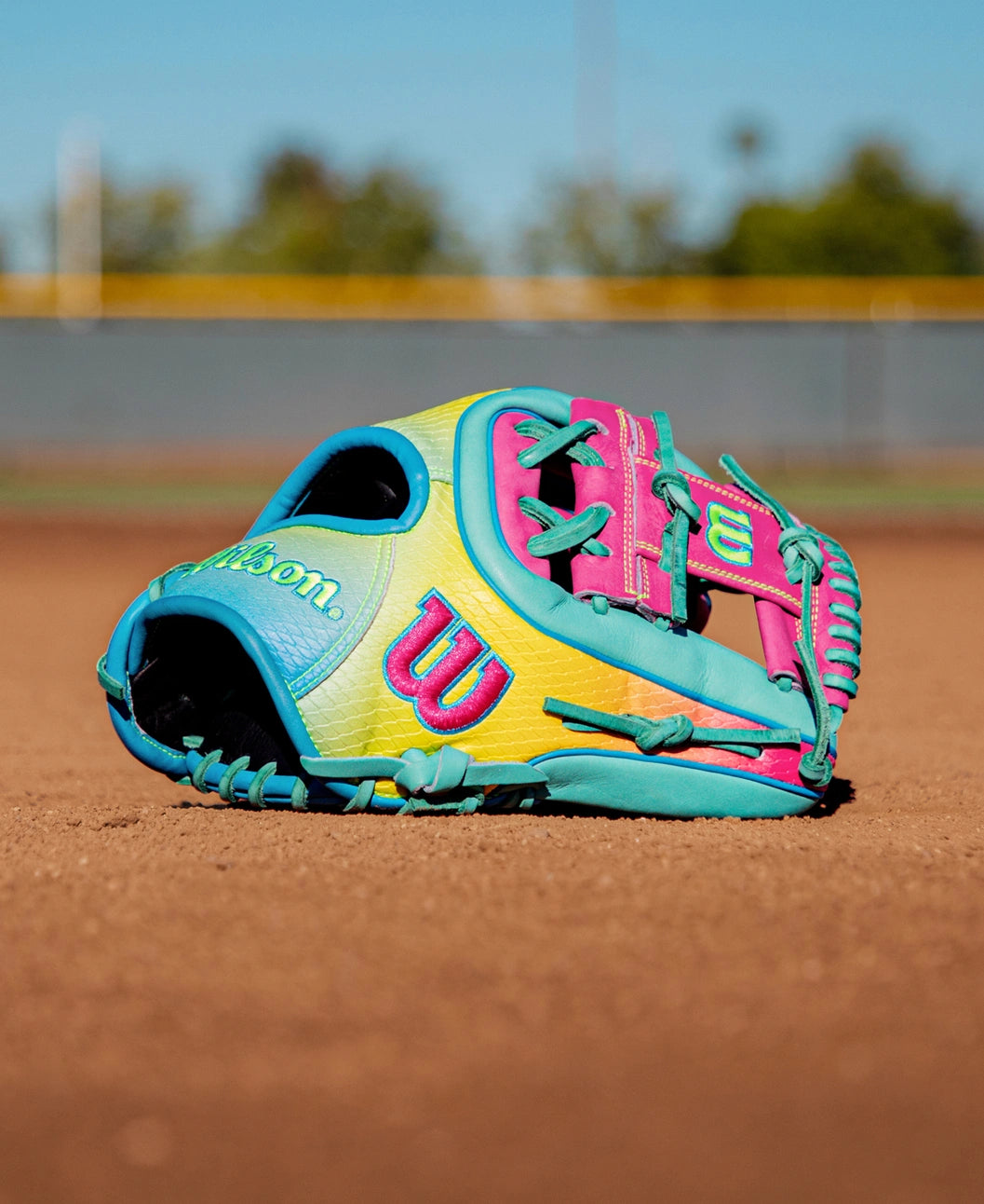 A Wilson A2000 1727SS 11.5″ Baseball Glove (WBG0009501RT1175) with eye-catching blue, pink, and yellow accents rests on a dirt baseball field, set against a backdrop of a blurred fence and trees. Crafted by Wilson with innovative SuperSnakeSkin material, this glove delivers vibrant style and top-tier performance for dedicated players.
