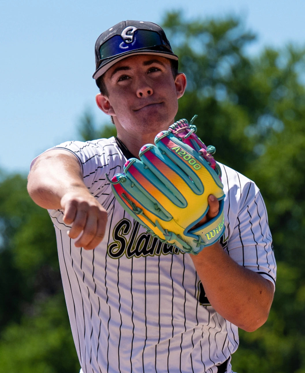 A baseball player in a pinstripe jersey throws a pitch while wearing the Wilson A2000 1727SS 11.5″ Baseball Glove (WBG0009501RT1175) featuring SuperSnakeSkin. Trees and blue sky are visible in the background.