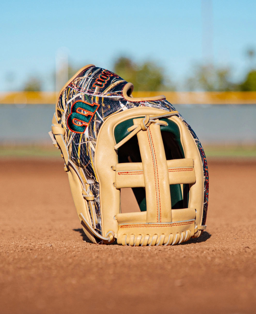 A Wilson A2000 GH2 11.75″ Gunnar Henderson GM Baseball Glove (WBG0009601RT1175) by Wilson, featuring tan leather with a camouflage pattern, sits upright on the dirt of a baseball field. In the background, a blurred fence and trees are visible beneath a clear blue sky.
