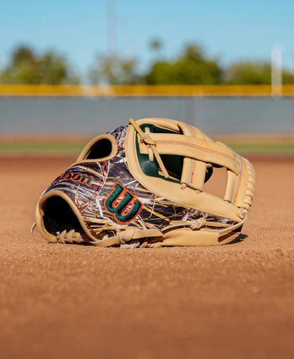 A Wilson A2000 GH2 11.75″ Gunnar Henderson GM Baseball Glove (WBG0009601RT1175) with a camouflage pattern lies on the dirt of an empty baseball field, with blurred green trees and a fence in the background.