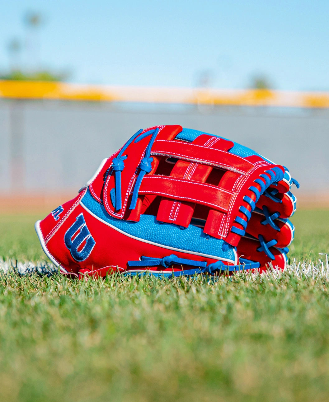 A Wilson A2000 PCA4 12.5″ Pete Crow-Armstrong GM Baseball Glove (WBG0009701) rests on green grass near a white line, with a blurred field and blue sky in the background.