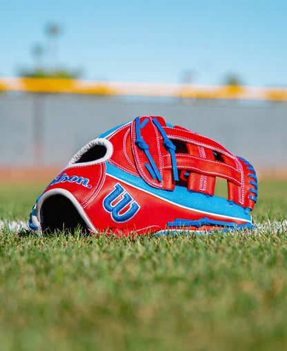 A Wilson A2000 PCA4 12.5″ Pete Crow-Armstrong GM Baseball Glove (WBG0009701) in red and blue lies on green grass near a white line, with a blurred field and sky in the background.