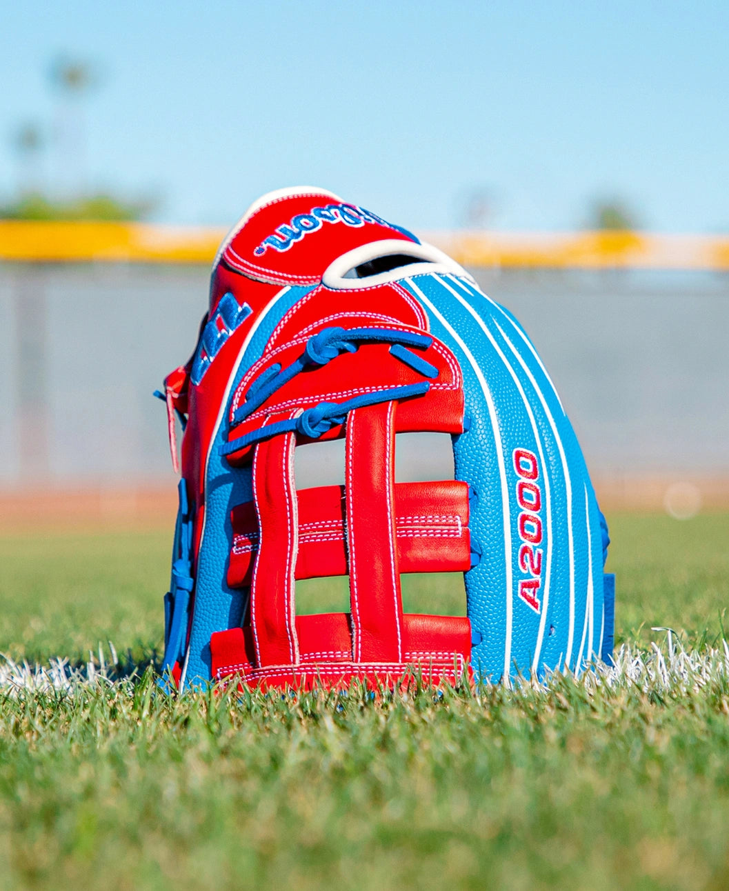 A Wilson A2000 PCA4 12.5″ Pete Crow-Armstrong GM Baseball Glove (WBG0009701) by Wilson, featuring red and blue accents, rests on green grass near a white line, with a blurred field and fence in the background.