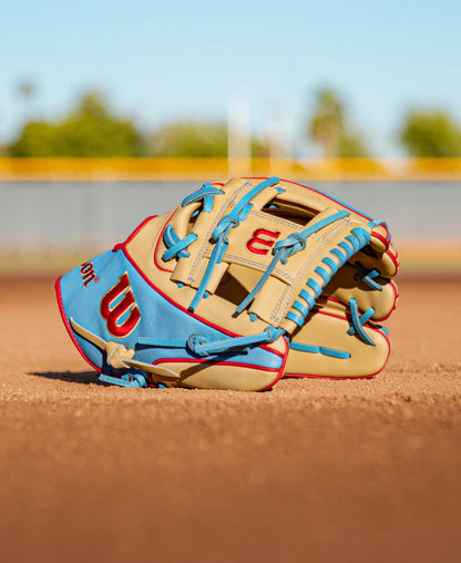 A Wilson A2K 1786 11.5″ Baseball Glove (WBG0010101RT115) in tan and blue Pro Stock Select leather, highlighted with bold red accents, rests on the dirt of a baseball field, with a blurred fence and green trees in the background.