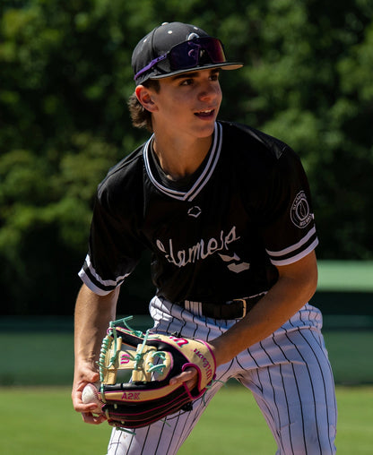 A baseball player in a black and white uniform prepares to throw a pitch on a sunny day, wearing sunglasses with trees in the background, and holding the Wilson A2K 1724 11.75″ Baseball Glove (WBG0010201RT1175) featuring Double Palm Construction.