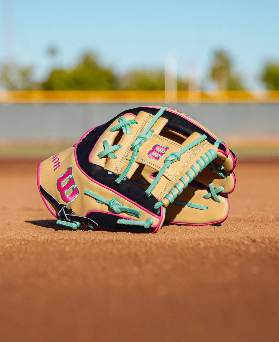 The Wilson A2K 1724 11.75″ Baseball Glove (WBG0010201RT1175) by Wilson, featuring a beige and navy design with teal laces and pink accents, lies on the dirt infield of a baseball field; its Double Palm Construction provides extra support, while grass and a fence blur gently in the background.