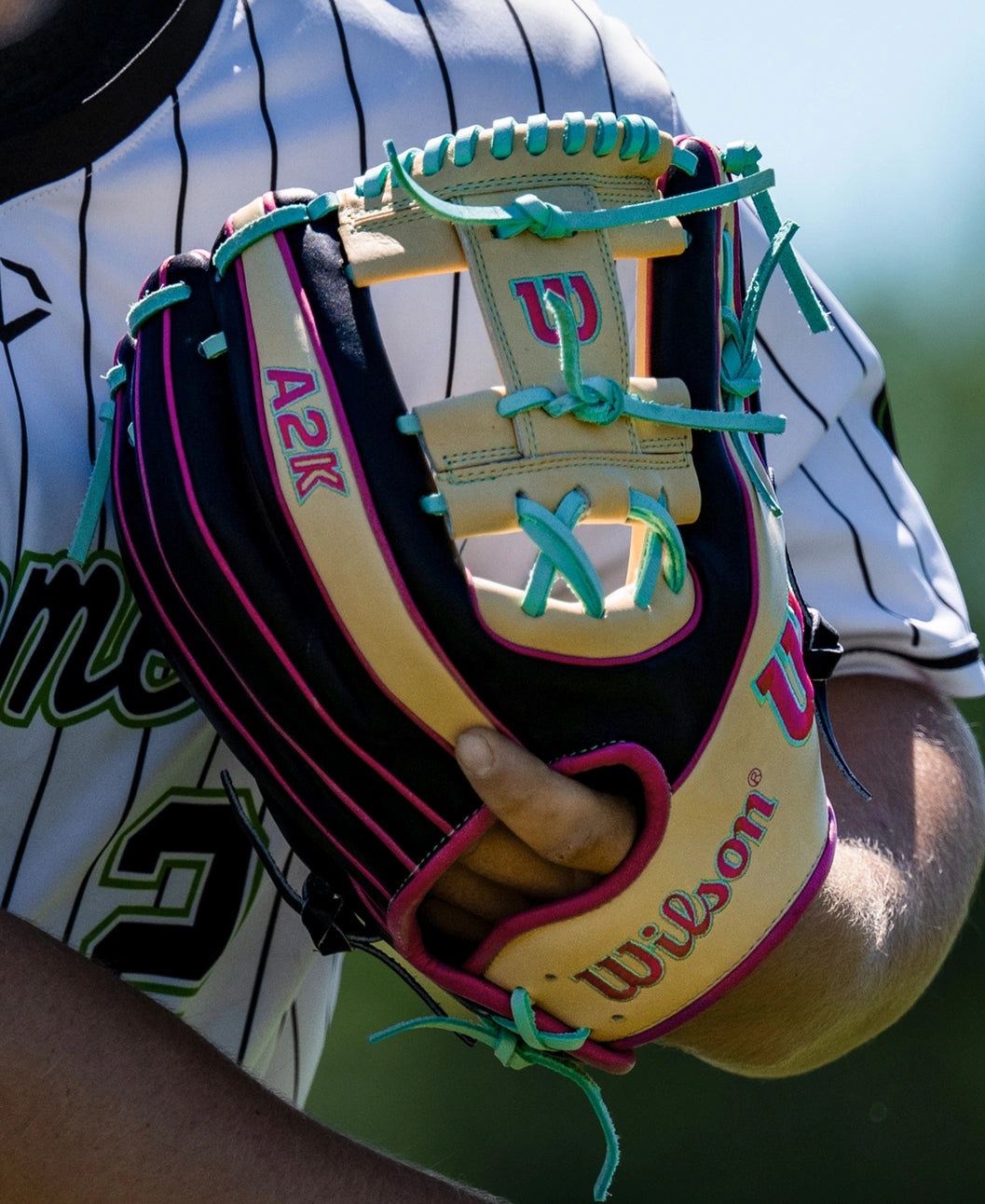 A close-up of a person wearing a striped baseball jersey holding the Wilson A2K 1724 11.75″ Baseball Glove (WBG0010201RT1175) with teal and pink laces, highlighting its Double Palm Construction for enhanced support.