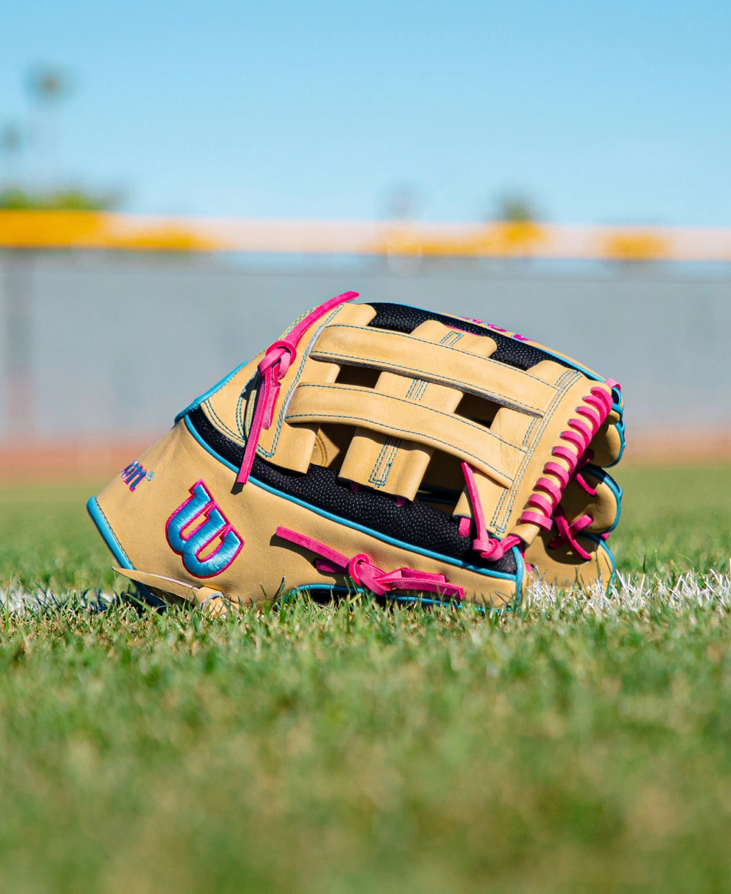 A Wilson A2K 1777 12.75″ Baseball Glove (WBG0010301) in tan and black with pink accents, featuring Double Palm Construction, rests on green grass against the backdrop of a blurred baseball field and a vibrant blue sky.