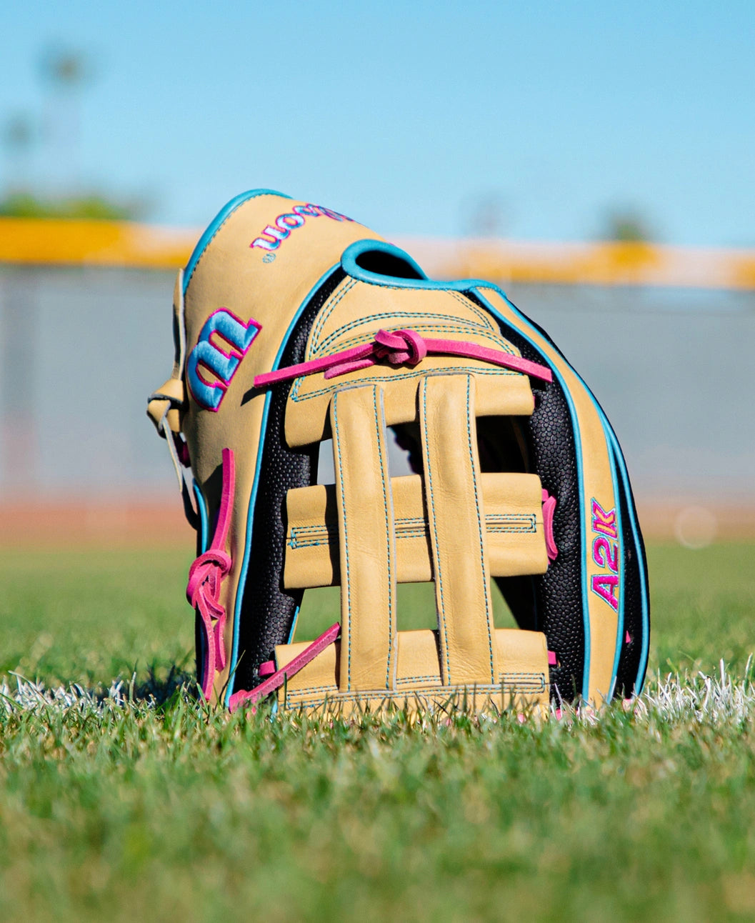 A Wilson A2K 1777 12.75″ Baseball Glove (WBG0010301) in tan with blue and pink accents stands upright on green grass, with a blurred baseball field and fence visible in the background under a clear blue sky, highlighting its Double Palm Construction for enhanced durability.