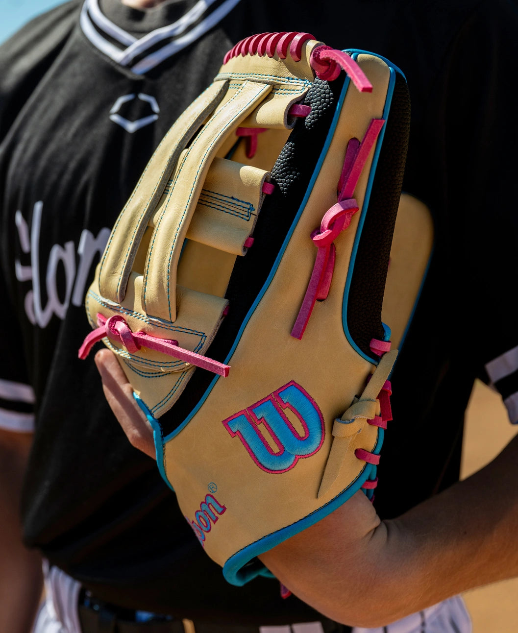 A person in a black baseball uniform holds a tan Wilson A2K 1777 12.75″ Baseball Glove (WBG0010301) by Wilson, featuring blue trim and pink laces.