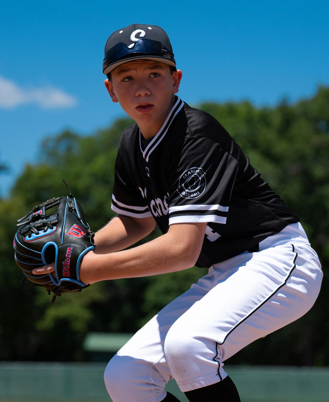 A young baseball player wearing a black and white uniform and a cap holds a black Wilson A1000 DP15 11.5″ Baseball Glove: WBG02402RTDP15 with blue trim in a ready position on a field.