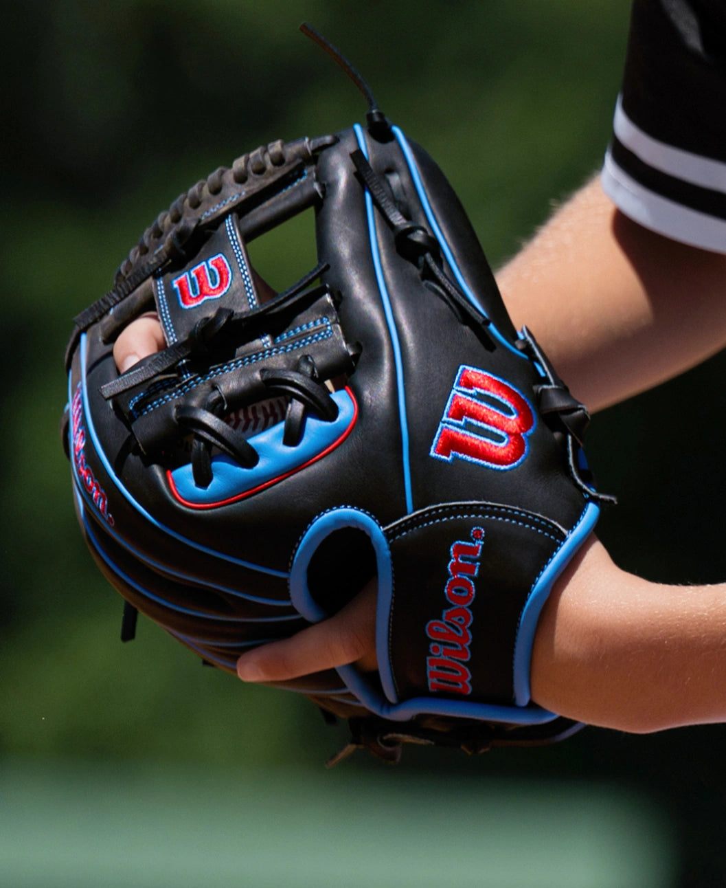 A player wearing a black Wilson A1000 DP15 11.5″ Baseball Glove: WBG02402RTDP15 with blue piping and red embroidered logos.