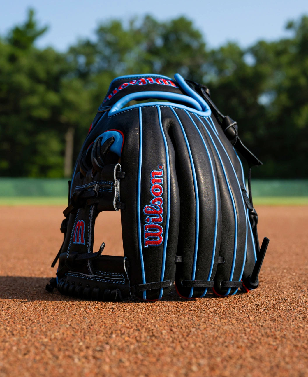 Wilson A1000 DP15 11.5″ Baseball Glove with black leather, blue piping, and red branding sitting on a dirt infield.