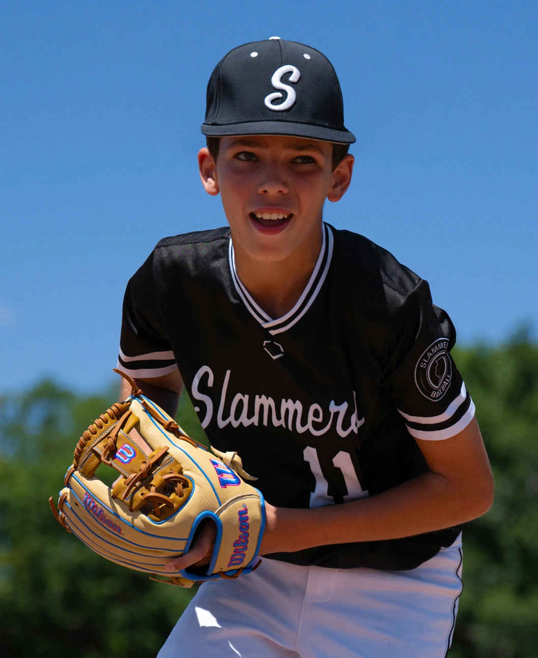A young baseball player in a black uniform and cap holds a Wilson A1000 1786 11.5″ Baseball Glove: WBG02501RT1786.