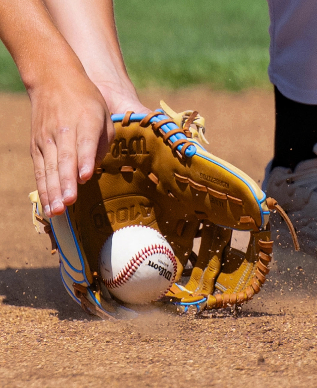 A baseball player uses a tan and light blue Wilson A1000 1786 11.5″ Baseball Glove: WBG02501RT1786 to field a ball rolling along a dirt infield.