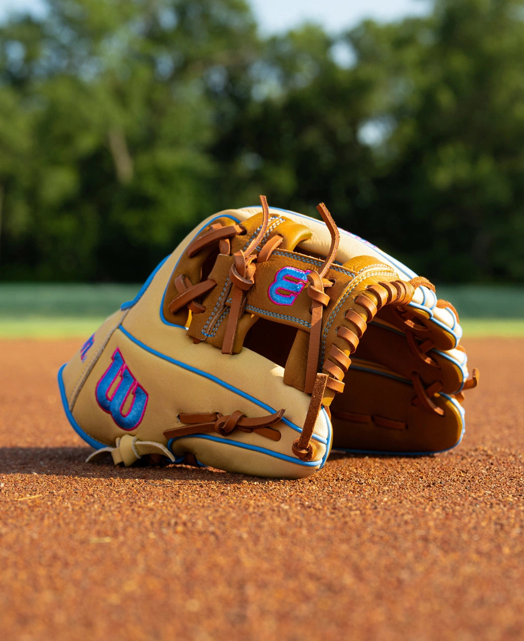 Wilson A1000 1786 11.5″ Baseball Glove with brown leather lacing and blue and pink embroidered logos resting on a dirt field.
