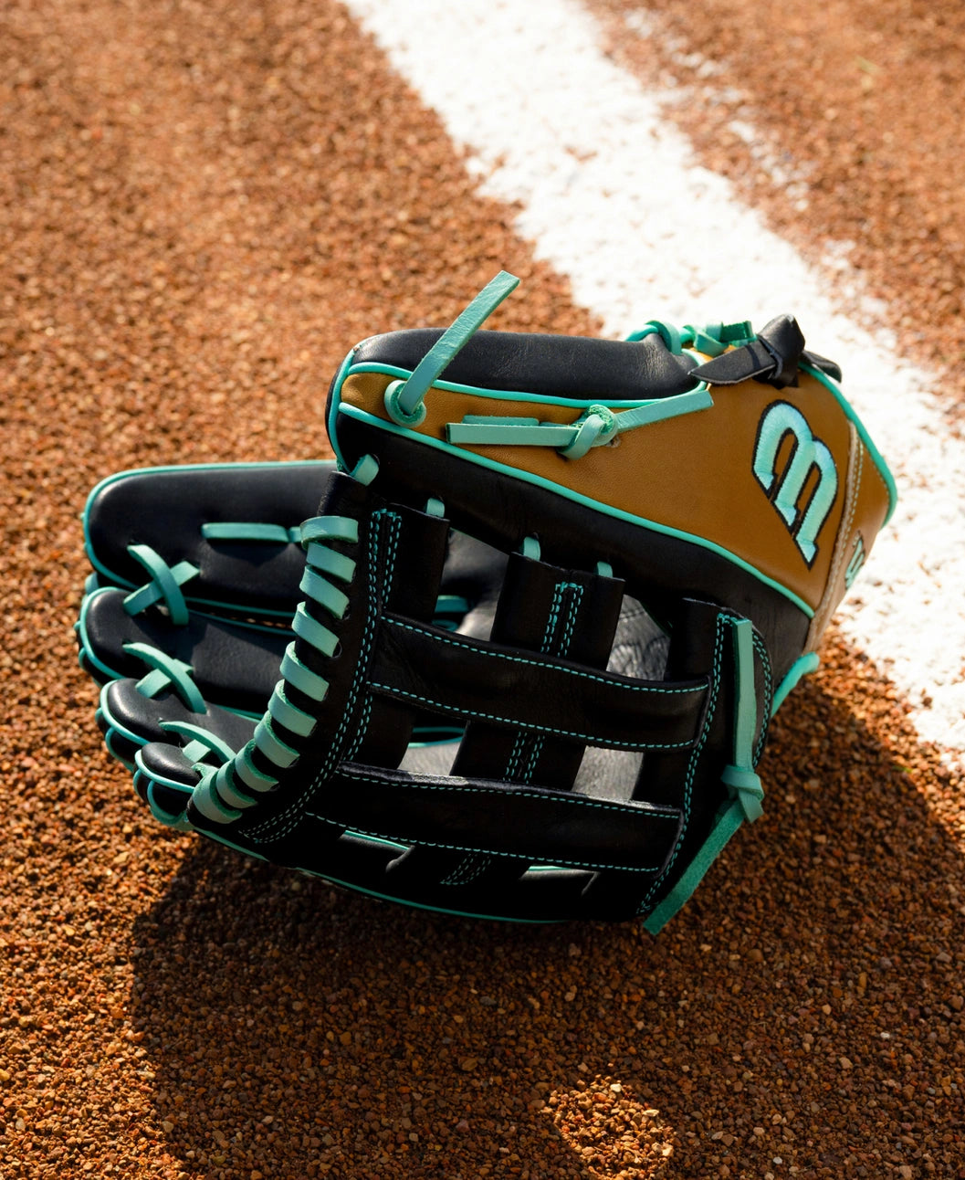 A black and brown Wilson A1000 DW5 12″ Baseball Glove: WBG02701RTDW5 with teal lacing resting on a dirt field.